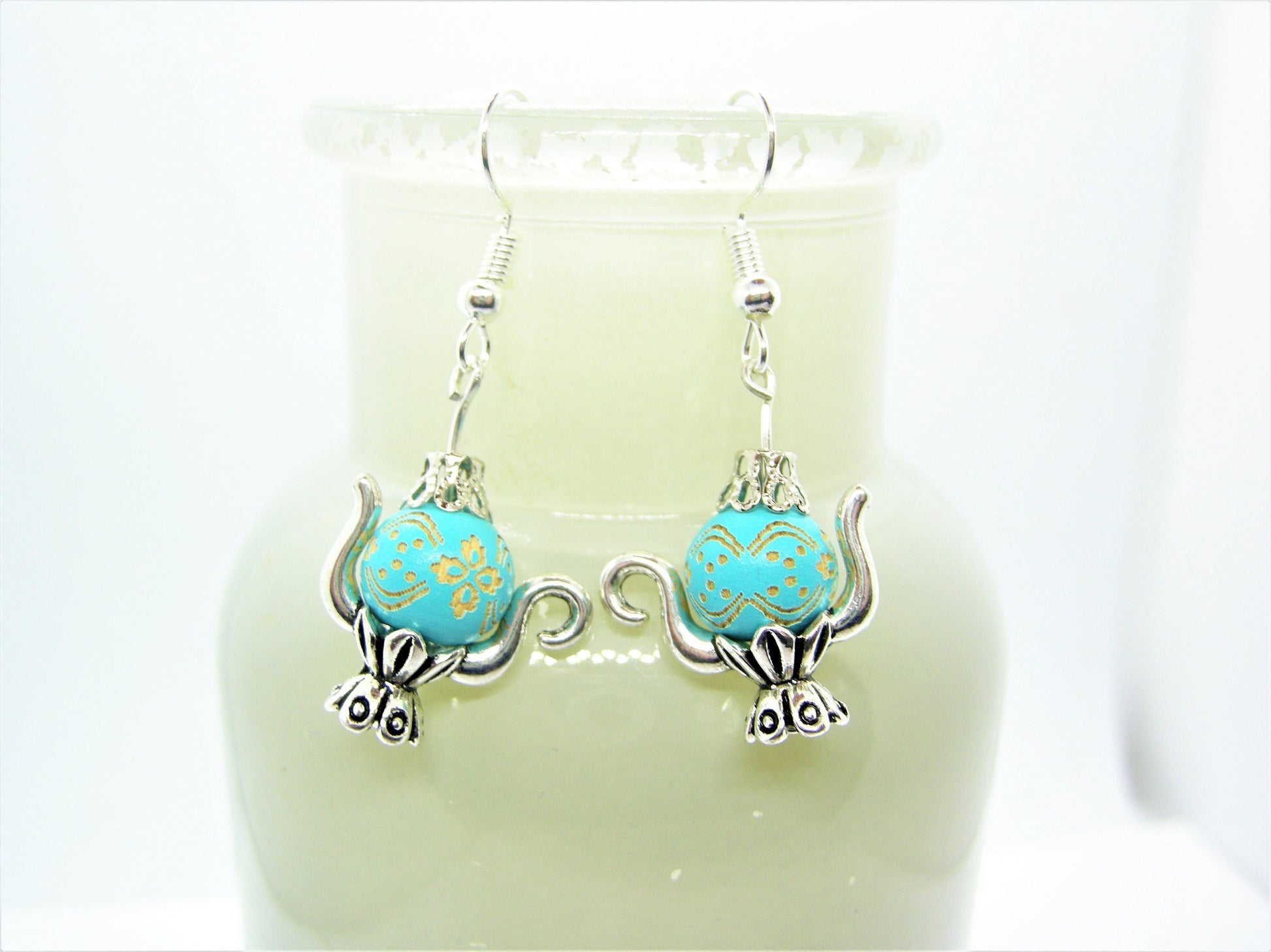 A pair of teapot-shaped earrings with blue and gold patterns, displayed on a white stand.